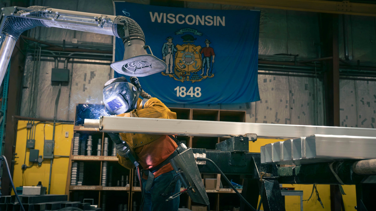A WiLL welder fabricating a light pole beneath a Wisconsin flag